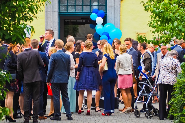 svatebčané se sešli před budovou, hodně jich stojí zády, muži v oblecích, ženy v šatech nebo sukních, modré balónky v pozadí